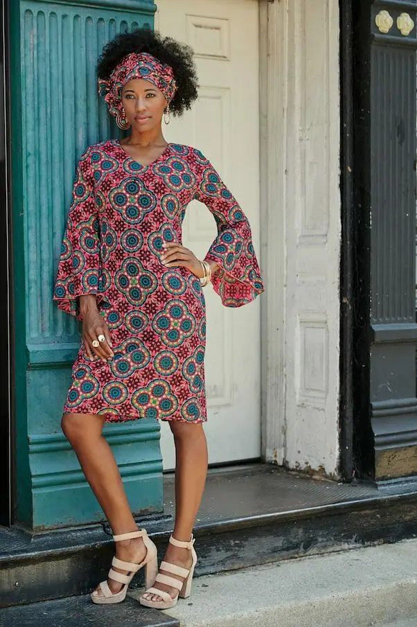 black woman wearing pink multi-print dress with flounce sleeves leaning against building