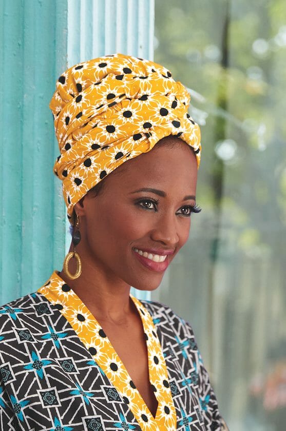 A smiling black woman wearing a yellow, black and white sunburst print headwrap, and a matching Afrocentric print top.