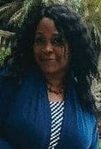 Ashro lookbook customer Karen, a black woman with long brown hair, in a blue jacket and print dress, with a red bag.