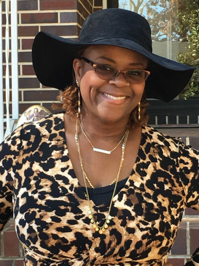 A smiling black woman with glasses, wearing a wide brimmed black hat and a leopard print top.