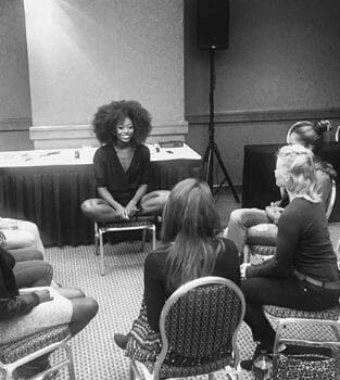 A black woman in an Afro, sitting crossed-legged with other women in a discussion circle.