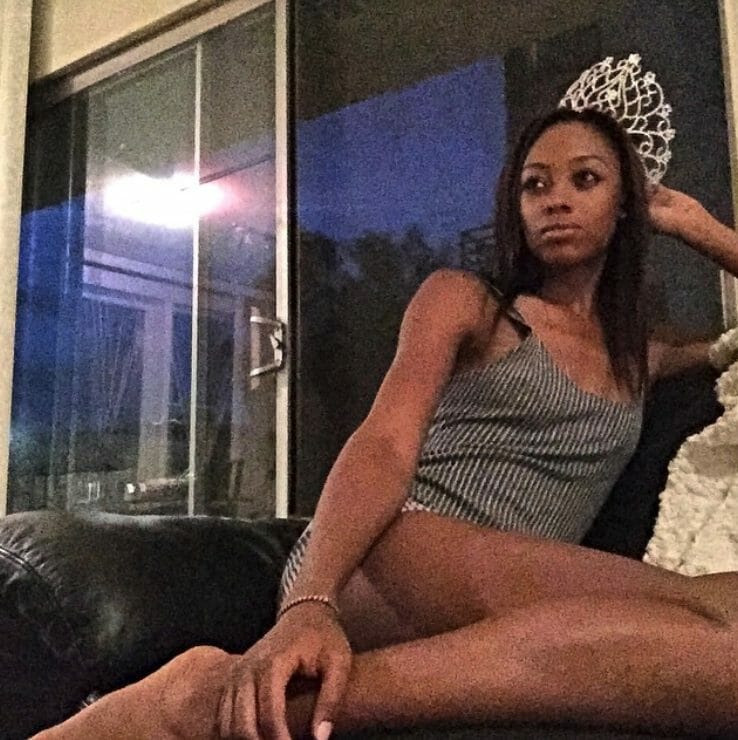 A black woman with long wavy hair, sitting on a couch, wearing a gray tank top and shorts.