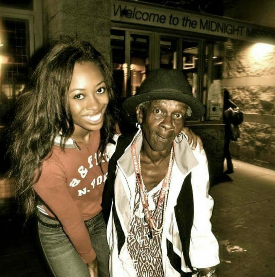 A smiling black woman with long hair, leaning over with her arm around an older black woman, in front of a building.
