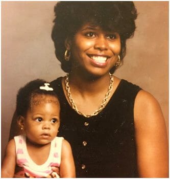 A smiling black woman in a black sleeveless top holding her little baby girl who has a bow in her hair.