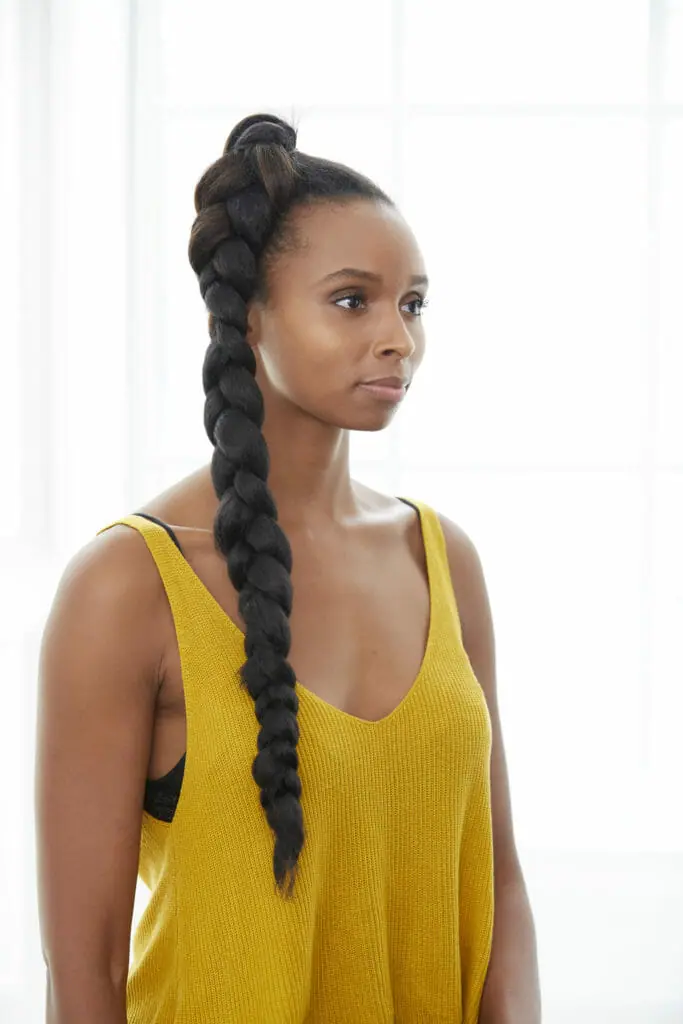 A black woman in a yellow sleeveless top, with a long braid at the top of her head, falling down to her chest.