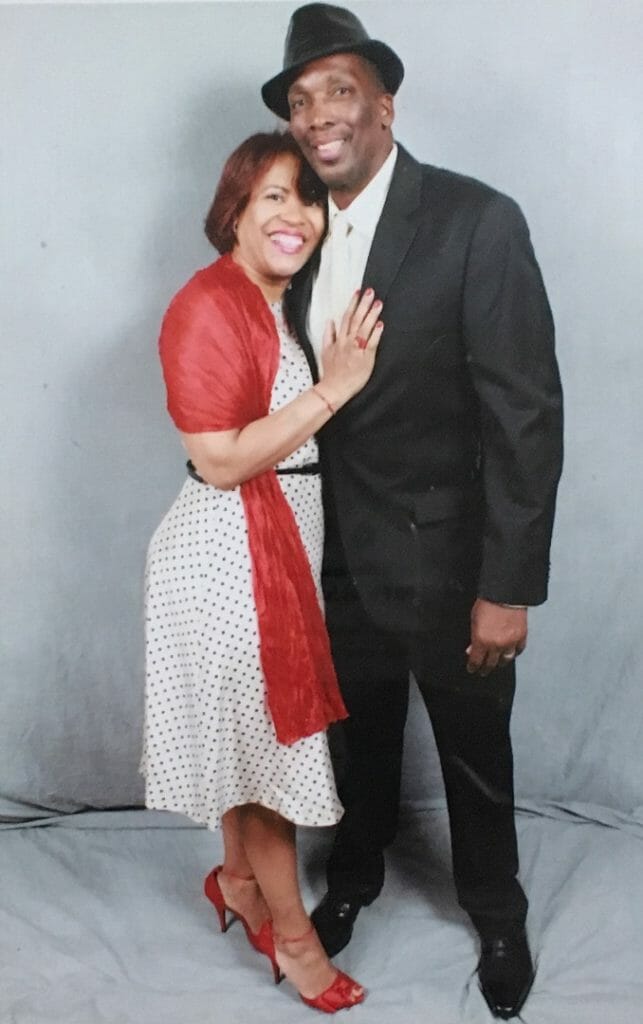 A black couple, with the woman in a white and black polka dot dress and red wrap, and the man in a black suit.