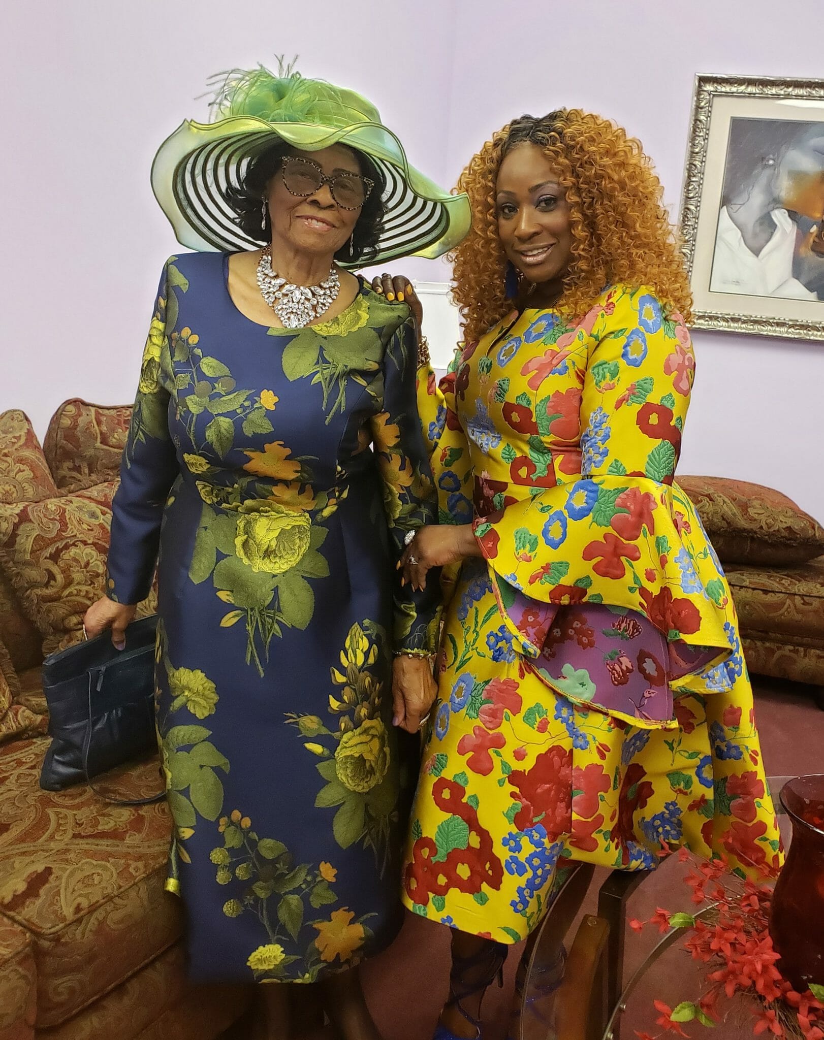 Ashro Woman of the Week, Alysha, a black woman with curly auburn hair, in a yellow floral dress with full skirt, and friend.