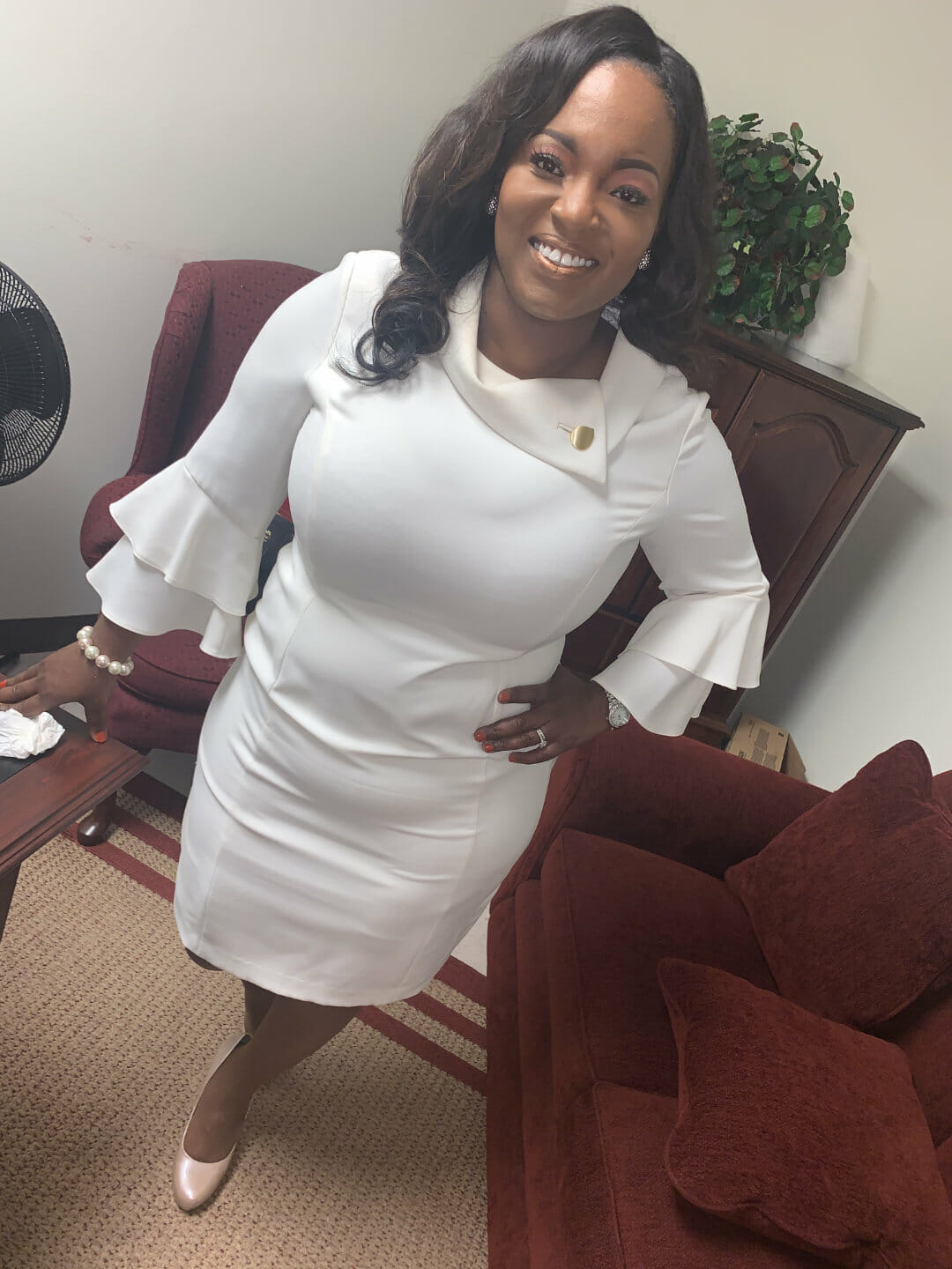 Ashro Woman of the Week ANGELA, a smiling black woman wearing a white dress, posed in her living room.