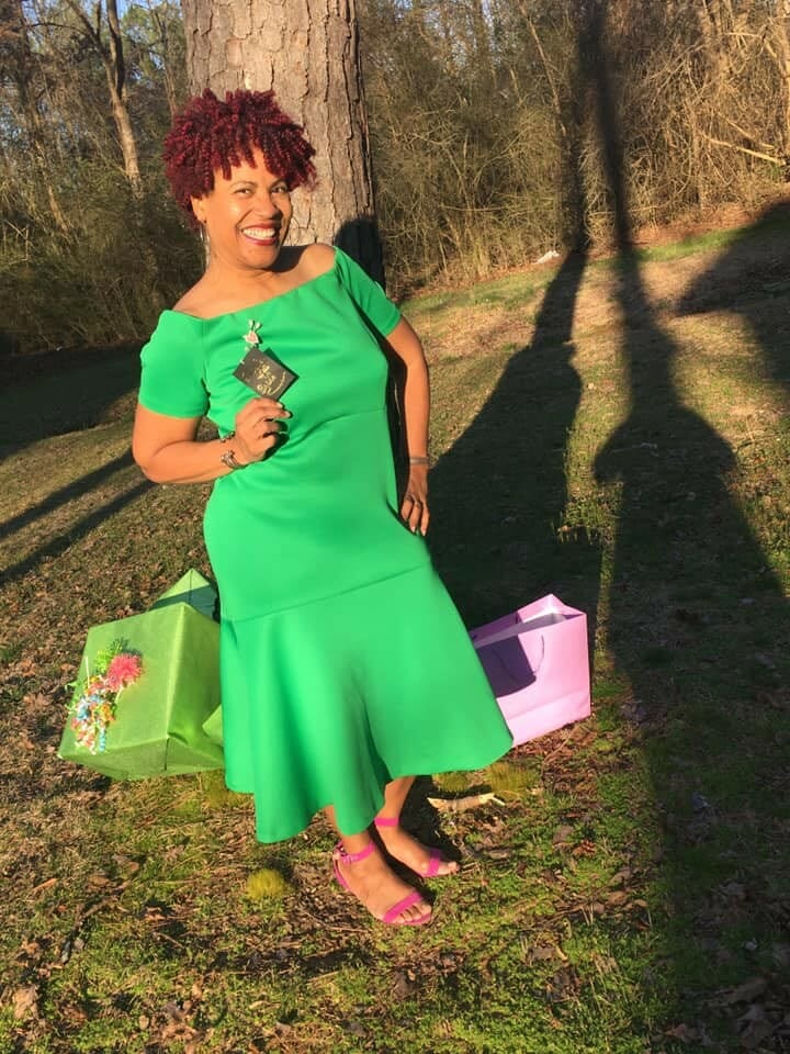 Leslie Royal smiling and wearing a gree dress standing by a tree and two shopping bags