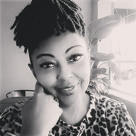 Smiling woman with locs resting her chin on her hand, wearing a leopard print top, with plants and sunlight in the background.