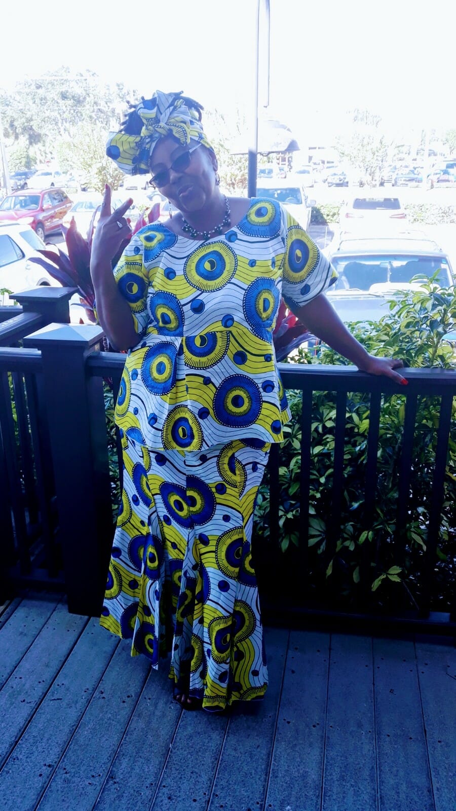 Woman in vibrant yellow and blue patterned outfit poses playfully outdoors, surrounded by greenery and parked cars.