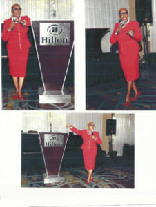 A woman in a red suit speaks confidently at a Hilton event, gesturing with enthusiasm at a podium.