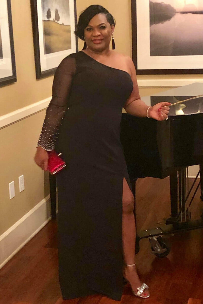 Woman in a black one-shoulder gown with a thigh-high slit, holding a red clutch, standing beside a piano in an elegant setting.