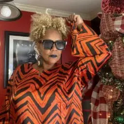 Woman posing in a orange and black pattern caftan with afrocentric earring and black sunglasses.
