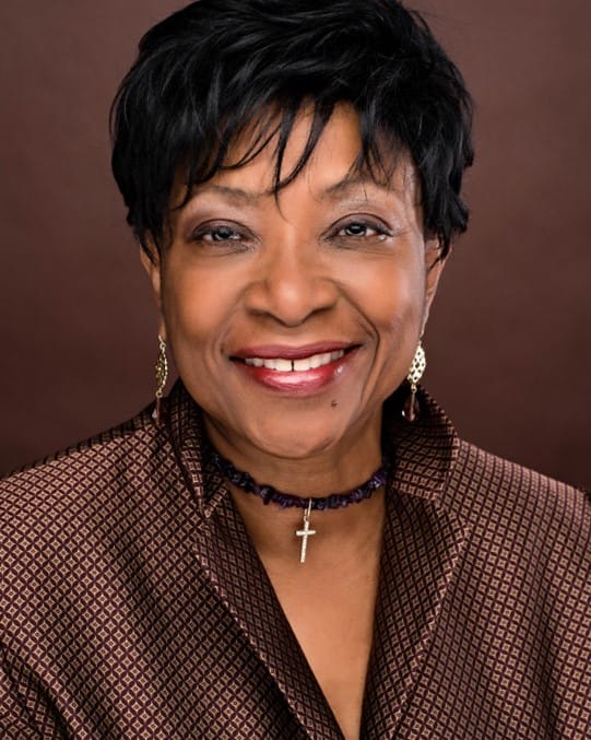 A smiling woman with short black hair wears a patterned brown jacket, gold earrings, and a purple beaded necklace with a cross pendant. She is posed against a plain brown studio background.