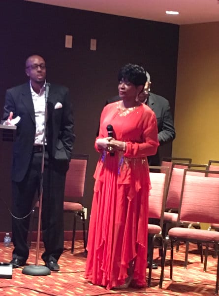 A woman in a bright red formal dress holds a microphone while standing in a carpeted room with rows of chairs. Two men in dark suits stand behind her, one speaking at a microphone stand.