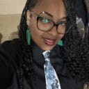 Woman with braided hair and glasses smiles, wearing a black shirt and a tie featuring historical figures