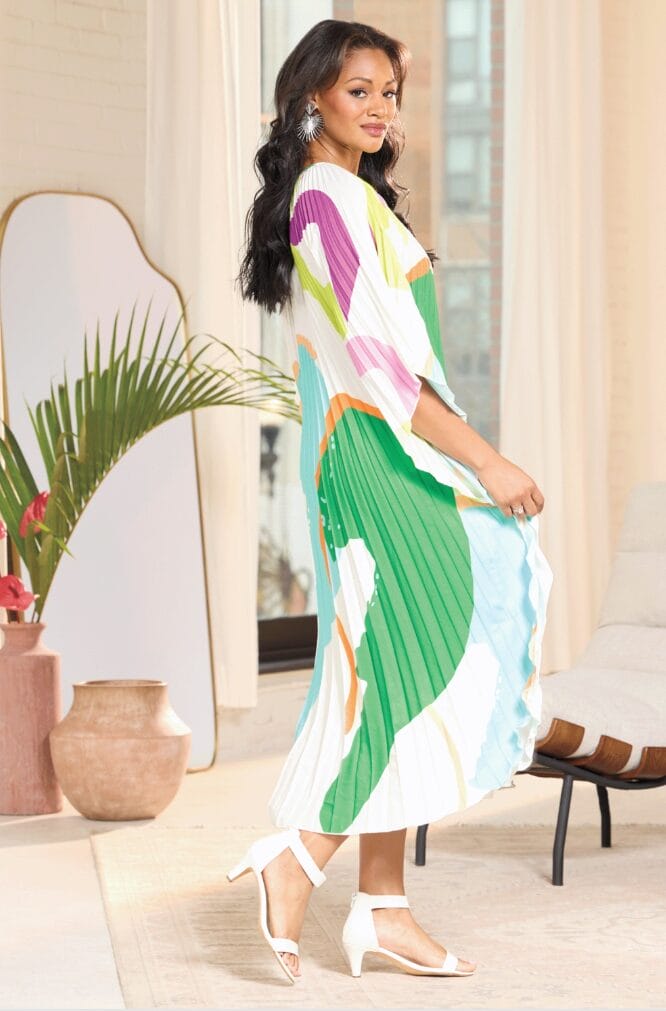 Model in a colorful, pleated dress poses gracefully, showcasing vibrant patterns and stylish white sandals.