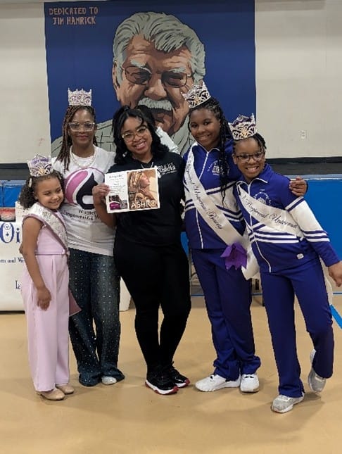 Five women and girls pose together, wearing crowns and sashes, holding a magazine, with a mural of a man in the background.
