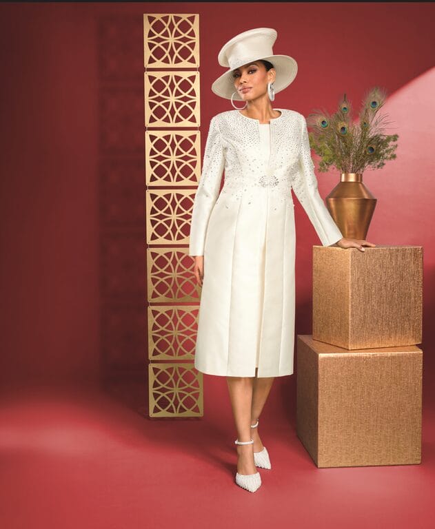 Woman in elegant cream suit and hat, adorned with rhinestones, stands near gold decorative blocks and peacock feathers.