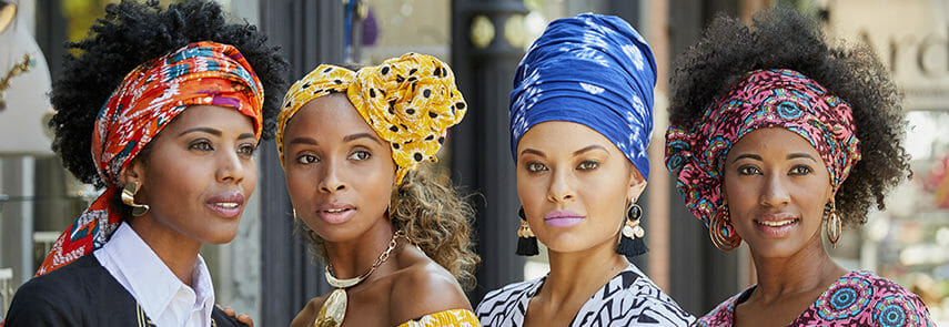 Four black women wearing different styles and prints of headwraps, in orange, yellow and black, blue, and pink and blue.