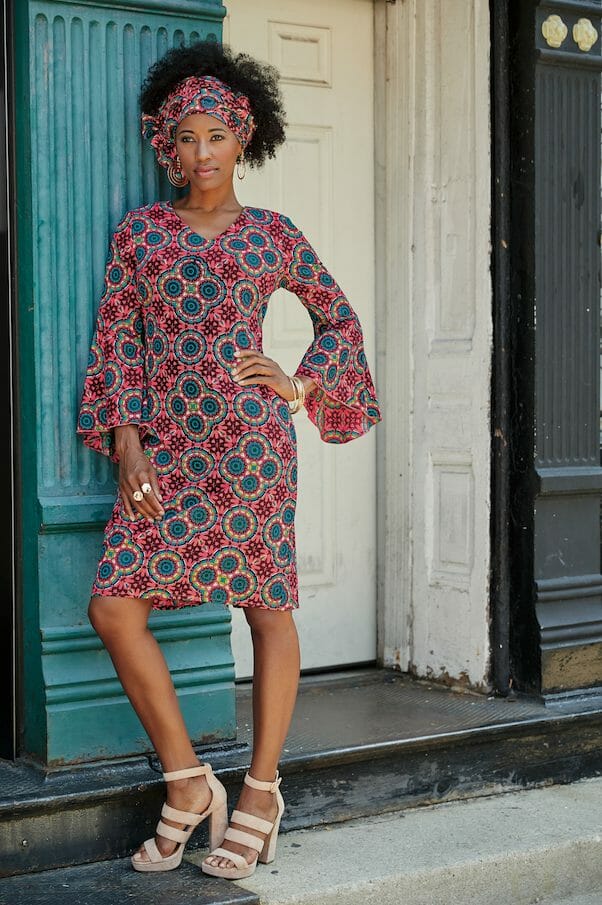 black woman wearing pink multi-print dress with flounce sleeves leaning against building