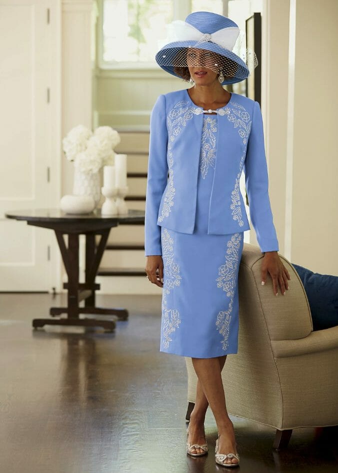 A black woman in a light blue jacket dress with white floral embroidery and a matching hat with a white band and netting.