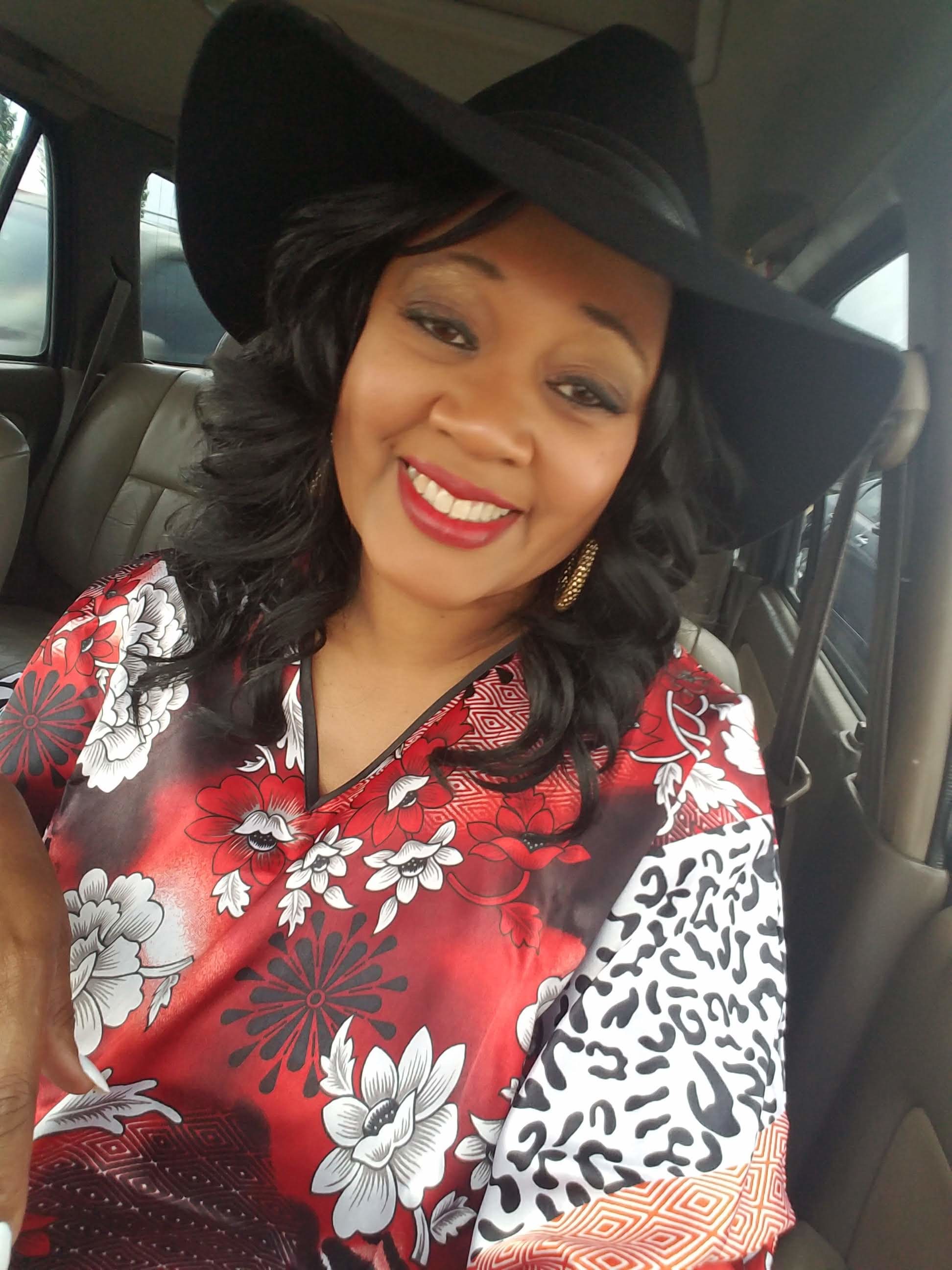 A black woman sitting in a car, wearing a black cowboy style hat and a red, white and black floral caftan.