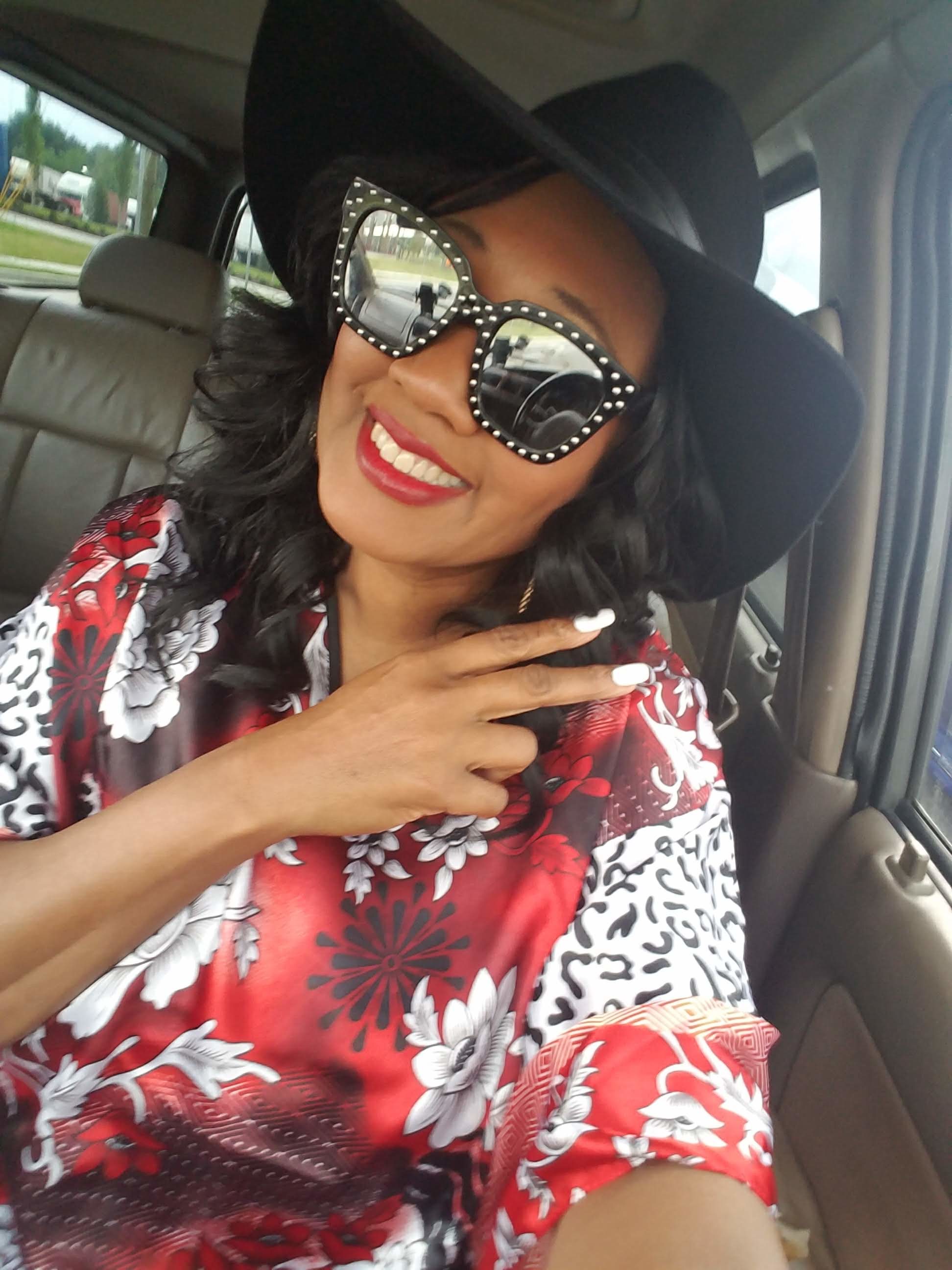 A black woman sitting in a car, wearing sunglasses and a black cowboy style hat and a red, white and black floral caftan.