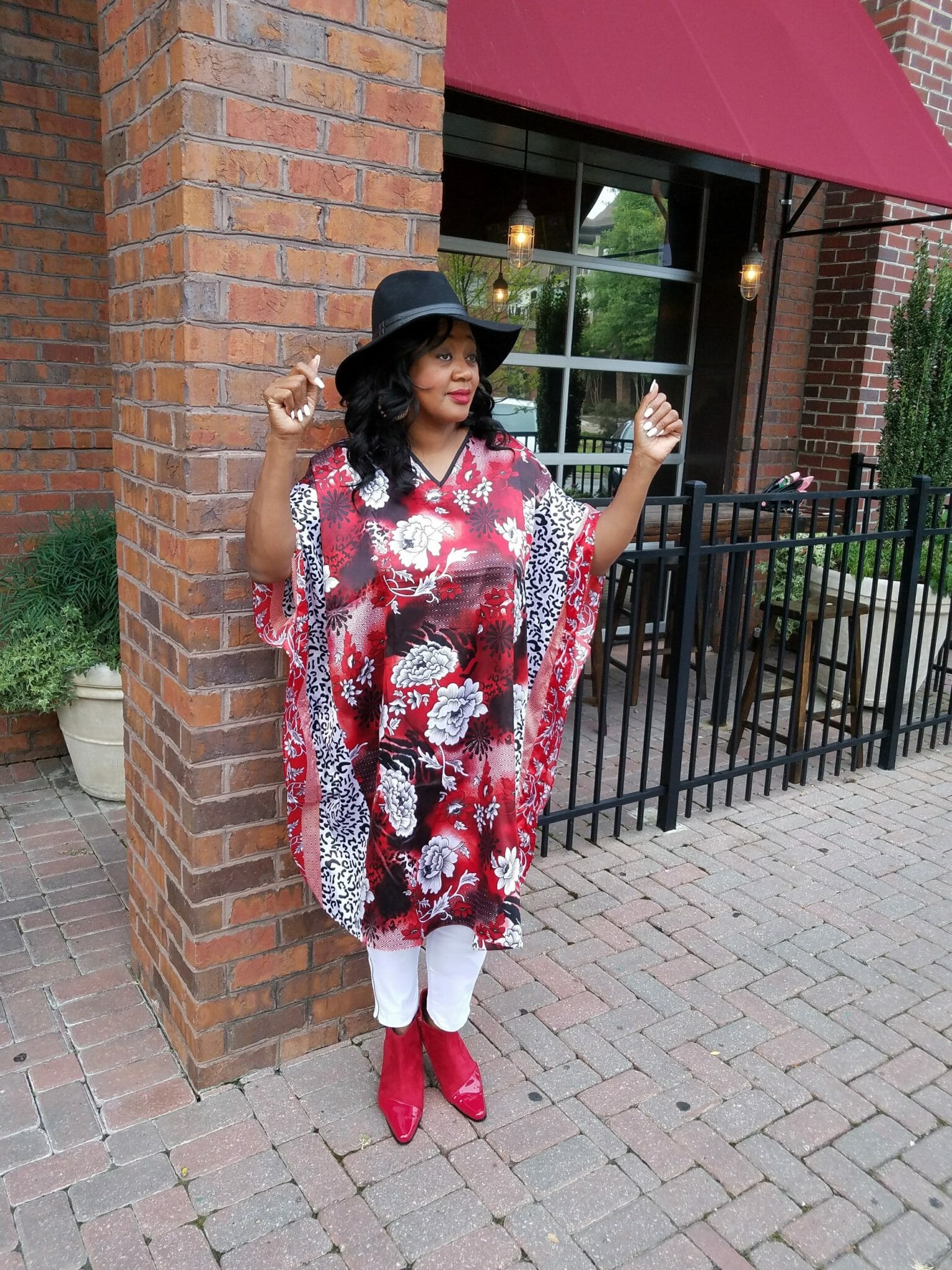 A black woman with arms up, wearing a black cowboy style hat and a red, white and black floral caftan and white pants.