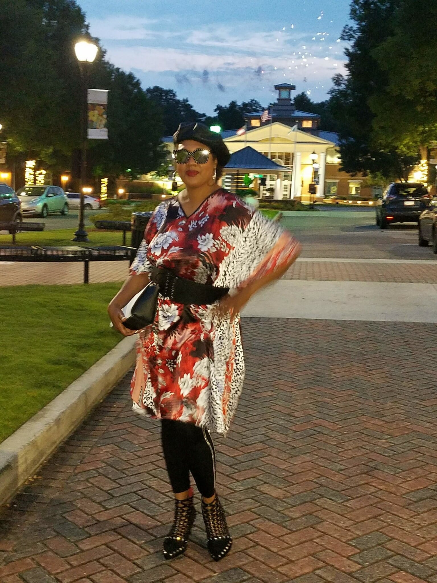 A black woman outside in a city at dusk, wearing a black beret and a red, white, and black floral caftan.