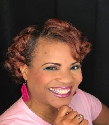 black woman with short wavy reddish hair smiling in pink shirt