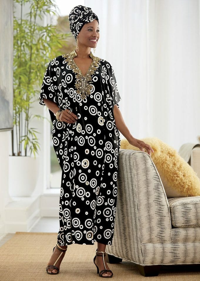 A smiling black woman wearing a long caftan in a black with white circles print and a matching headwrap.