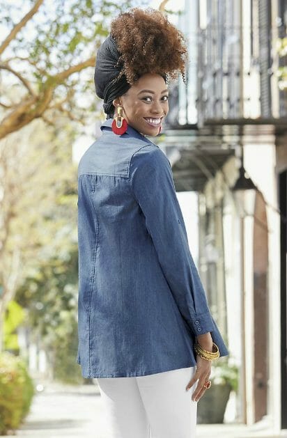 Back view of a smiling black woman wearing red earrings, a black headwrap, a blue denim tunic and white pants.