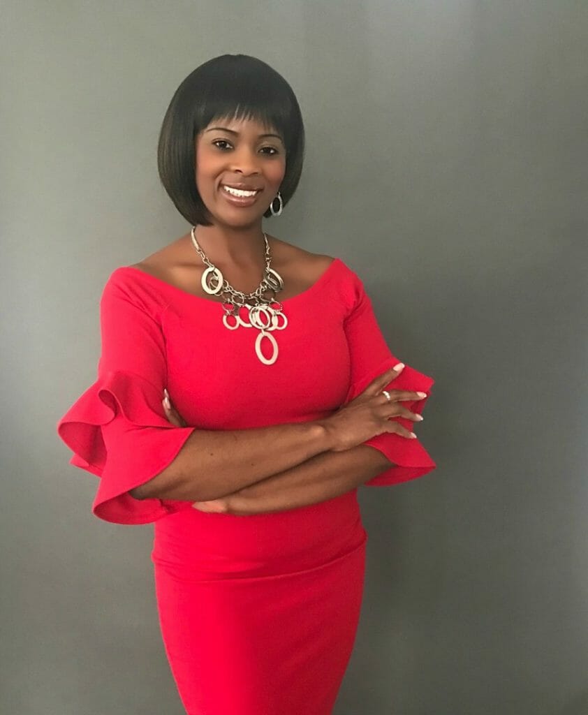 A smiling African-American woman wearing a red dress with flounced sleeves and a long, gold circle necklace.