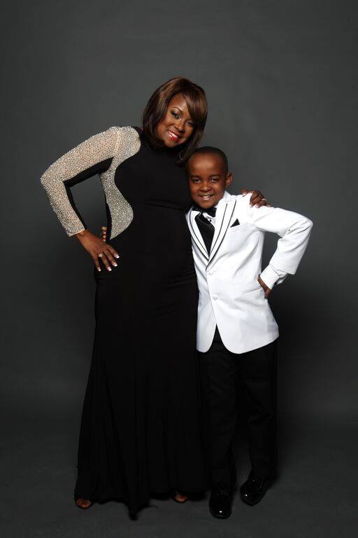 A smiling black woman in a black and gray dress, standing with a black boy in a white suit jacket and black pants.