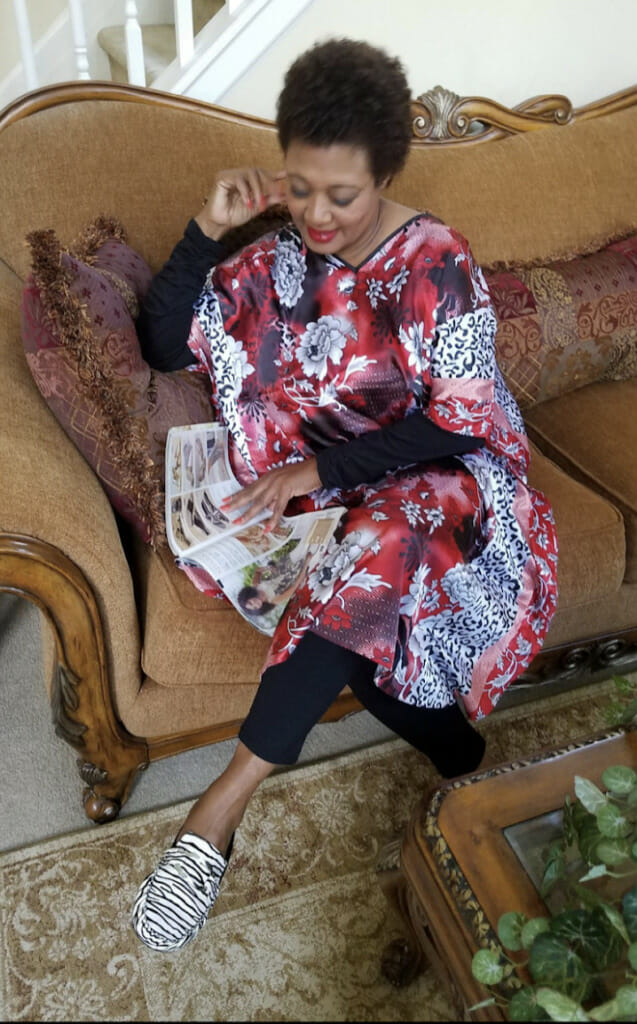 A black woman sitting on a brown chaise, wearing a red, white and black floral caftan with black leggings.