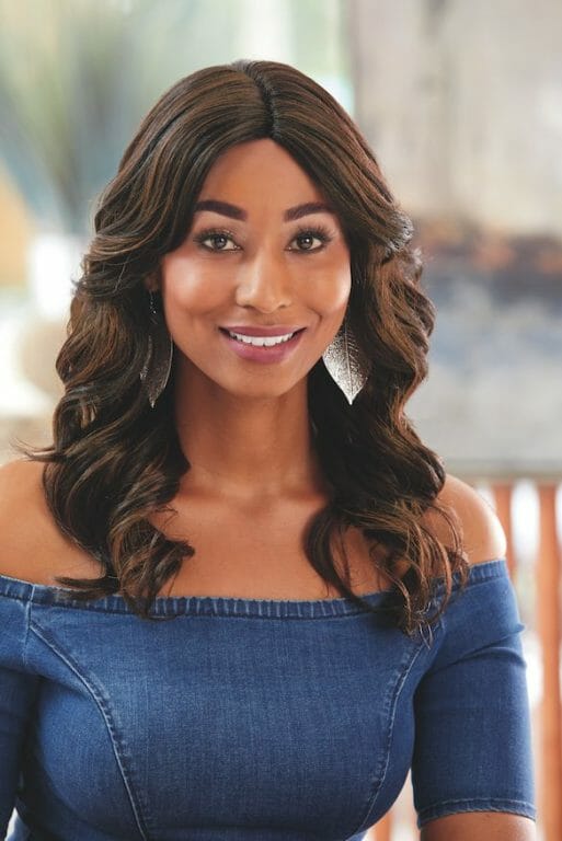 A smiling black woman in a long, brown wavy wig wearing a denim blue off-the-shoulder top.