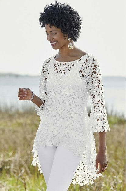 A black woman in a white floral cutout tunic, white cami and white pants at a lake shore.