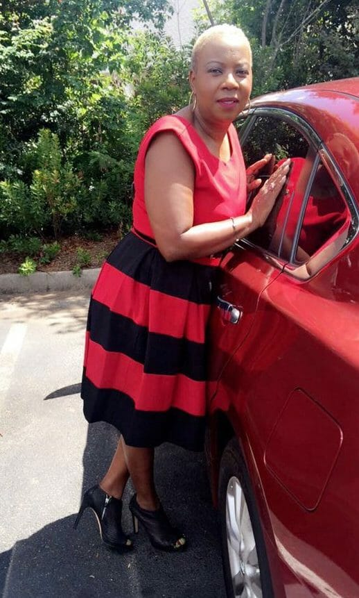 A black woman with short hair, wearing a red and black striped sleeveless dress, and leaning up against a red car.