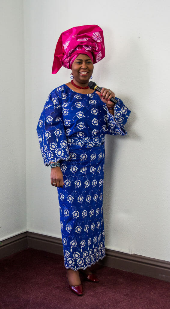 A black woman wearing a big pink headwrap and a long, blue and white Afrocentric print dress.
