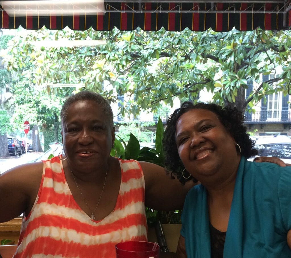 A black woman in a red and white sleeveless top sitting in a restaurant with a smiling black woman in a blue top.