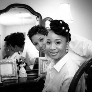 Two smiling African-American sisters, one in a fancy hairdo, seated in front of a mirrored vanity.