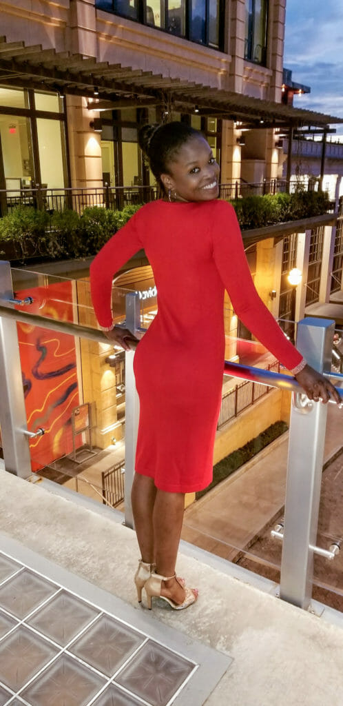 A smiling black woman in a fitted long-sleeve red dress, standing on a balcony, looking over her shoulder.