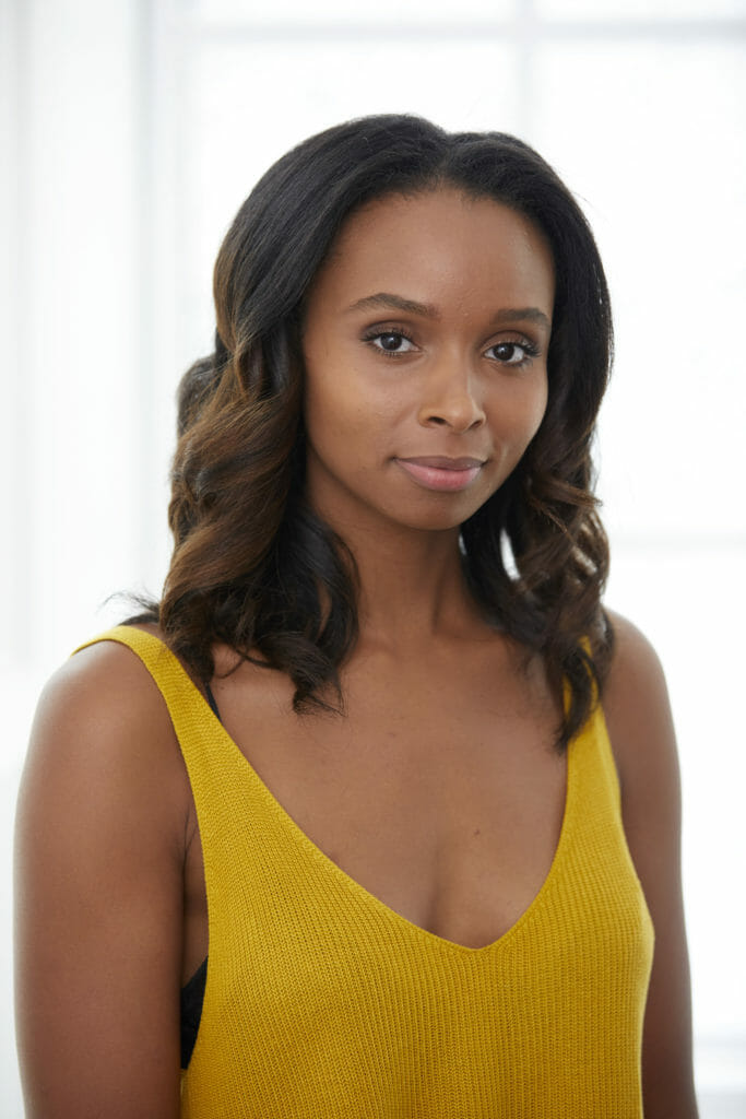 An African-American woman in a yellow sleeveless top, with wavy shoulder-length hair.