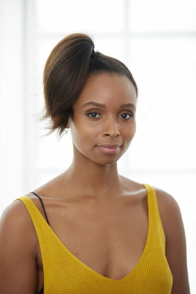 An African-American woman in a yellow sleeveless top, with a ponytail at the top of her head.