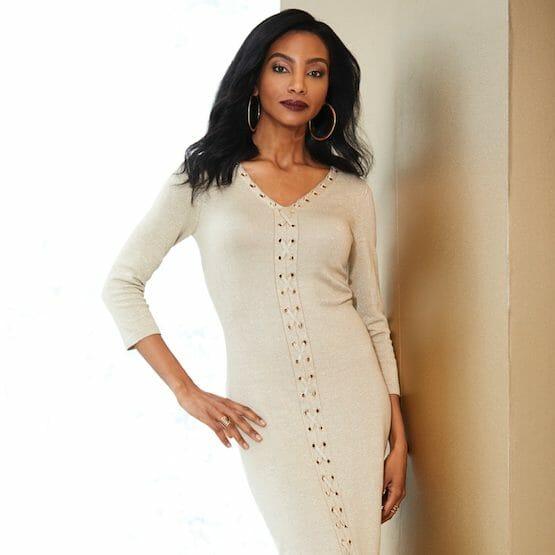 An African-American woman in a fitted beige V-neck dress with corset-like detail down the front to the hem.