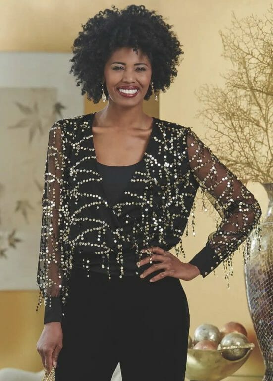A smiling African-American woman in a sheer black top with gold fringe sequin detail and black pants.