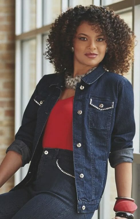 An African-American woman with dark brown cury hair, wearing a red cami with a denim jacket and jeans.