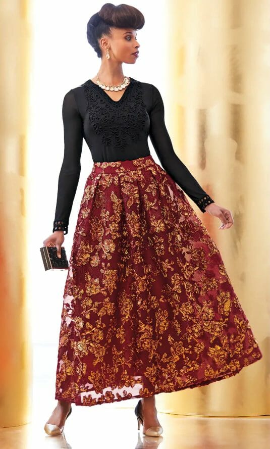 An African-American woman with a fancy updo hairstyle, wearing a black V-neck top, pearl necklace and a red floral skirt.